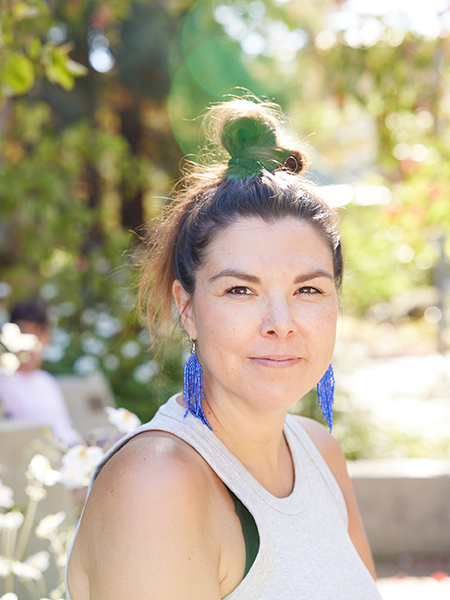 Asta Mønsted smiles and looks at the camera. The assistant professor has her hair in a bun atip her head with long blue beaded earrlings and a white tank top. She is outdoors.