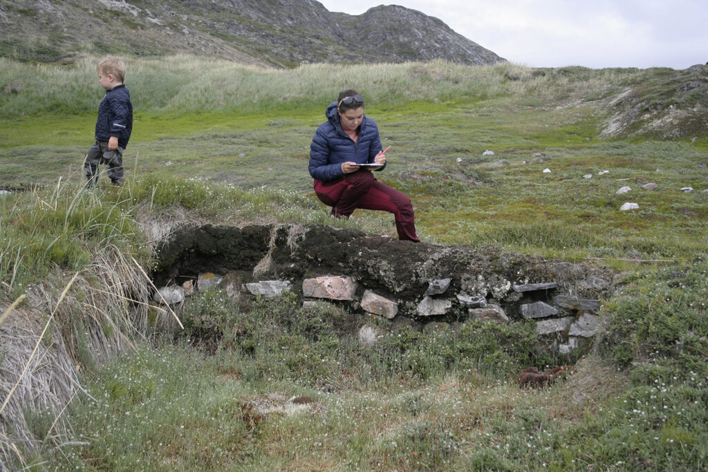 Assistant professor Asta Mønsted does fieldwork in Greenland, pausing to take notes while crouched in front of a mound with large rocks in it.