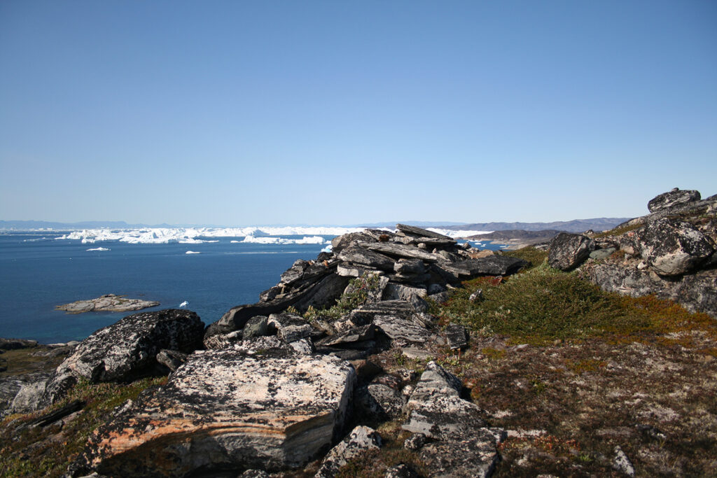 A rocky oceanside landscape in Greenland also shows icy mounds in the distance waters.