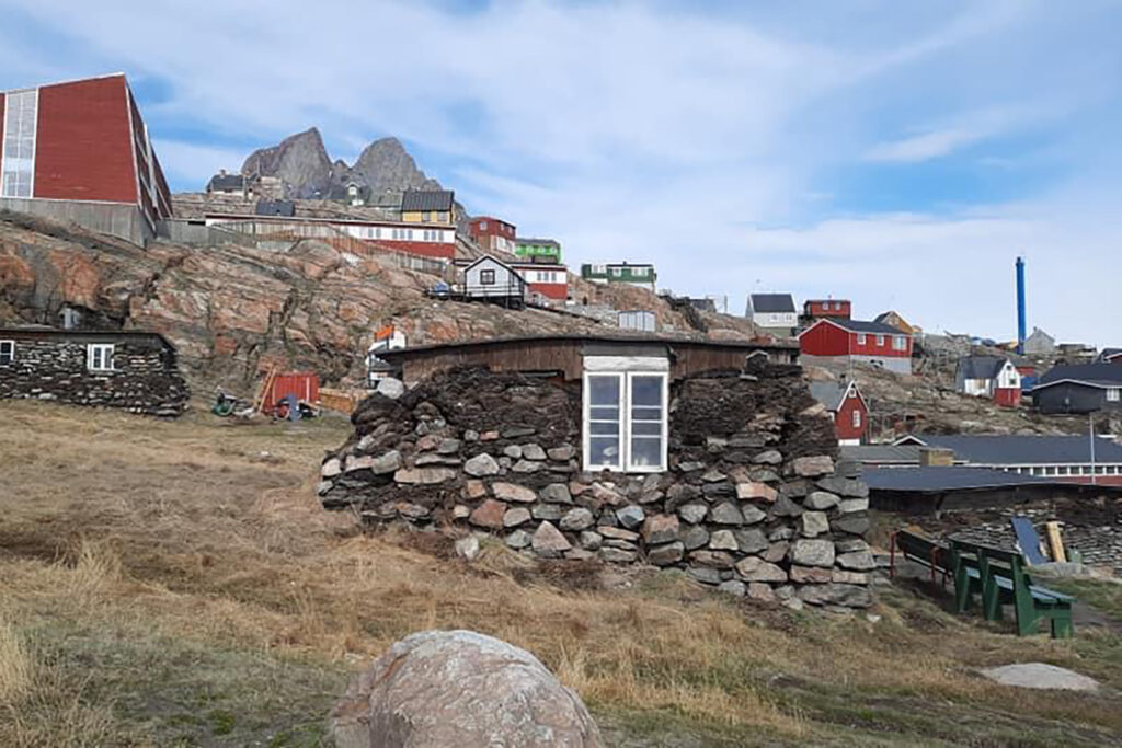 Three historic stone-and-turf houses in Greenland stand near a modern structure, the local school. 