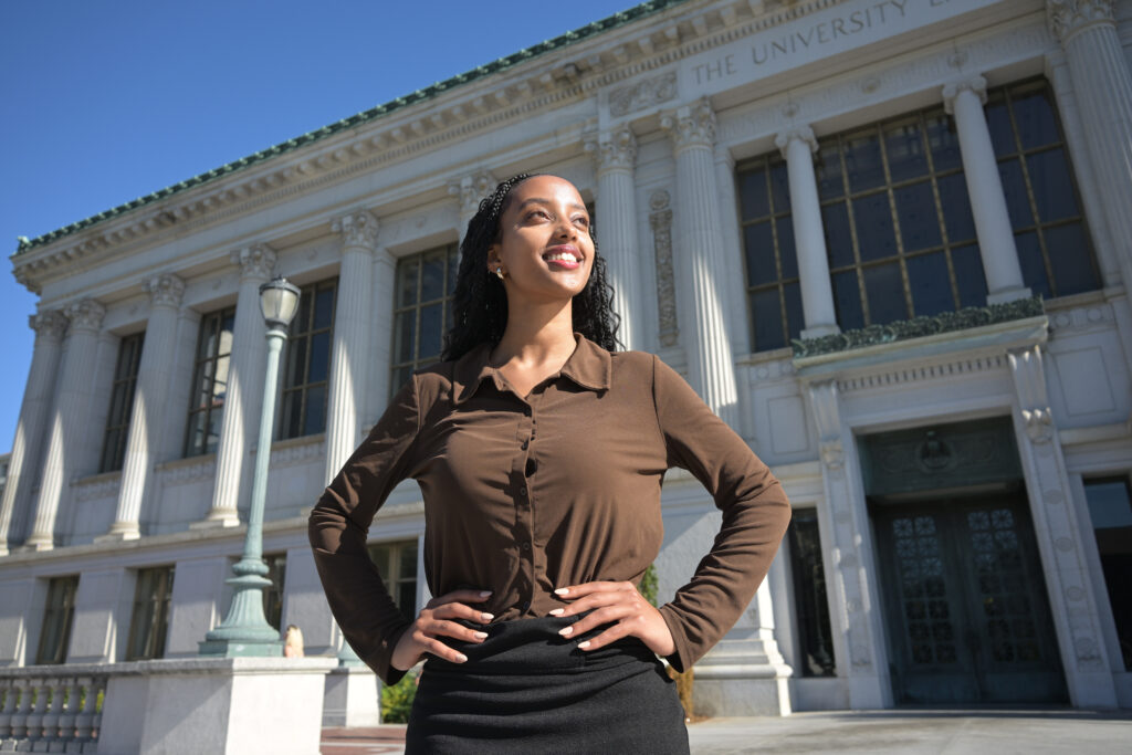 Beteliham Mamo, wearing brown shirt with her hands on her hips and smiling, looks proudly to the side while standing in front of the library at UC Berkeley.