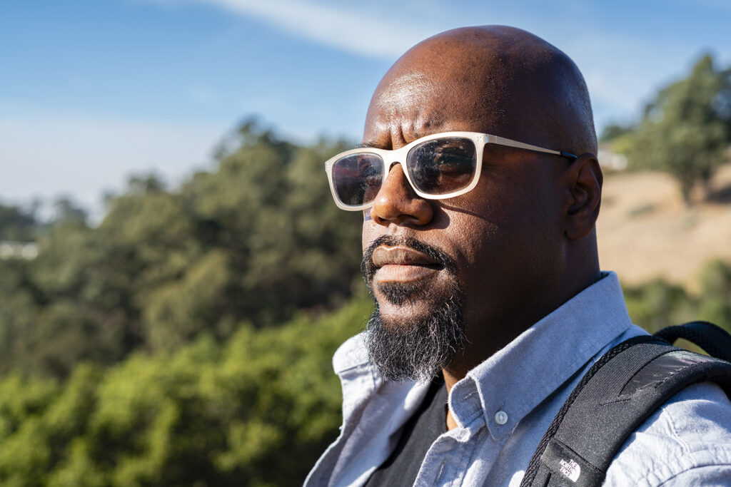 Professor Chris Schell wears white-rimmed sunglasses and looks out into the distance. He is African American and has a beard, mustache and a collared shirt on. He is out in nature.