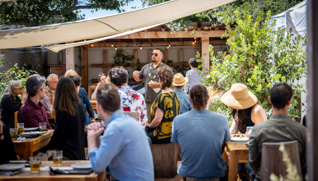 A crowd of diners sit at wooden tables on a patio that is partially covered by a white cloth awning. A person is standing among the diners and speaking to them.