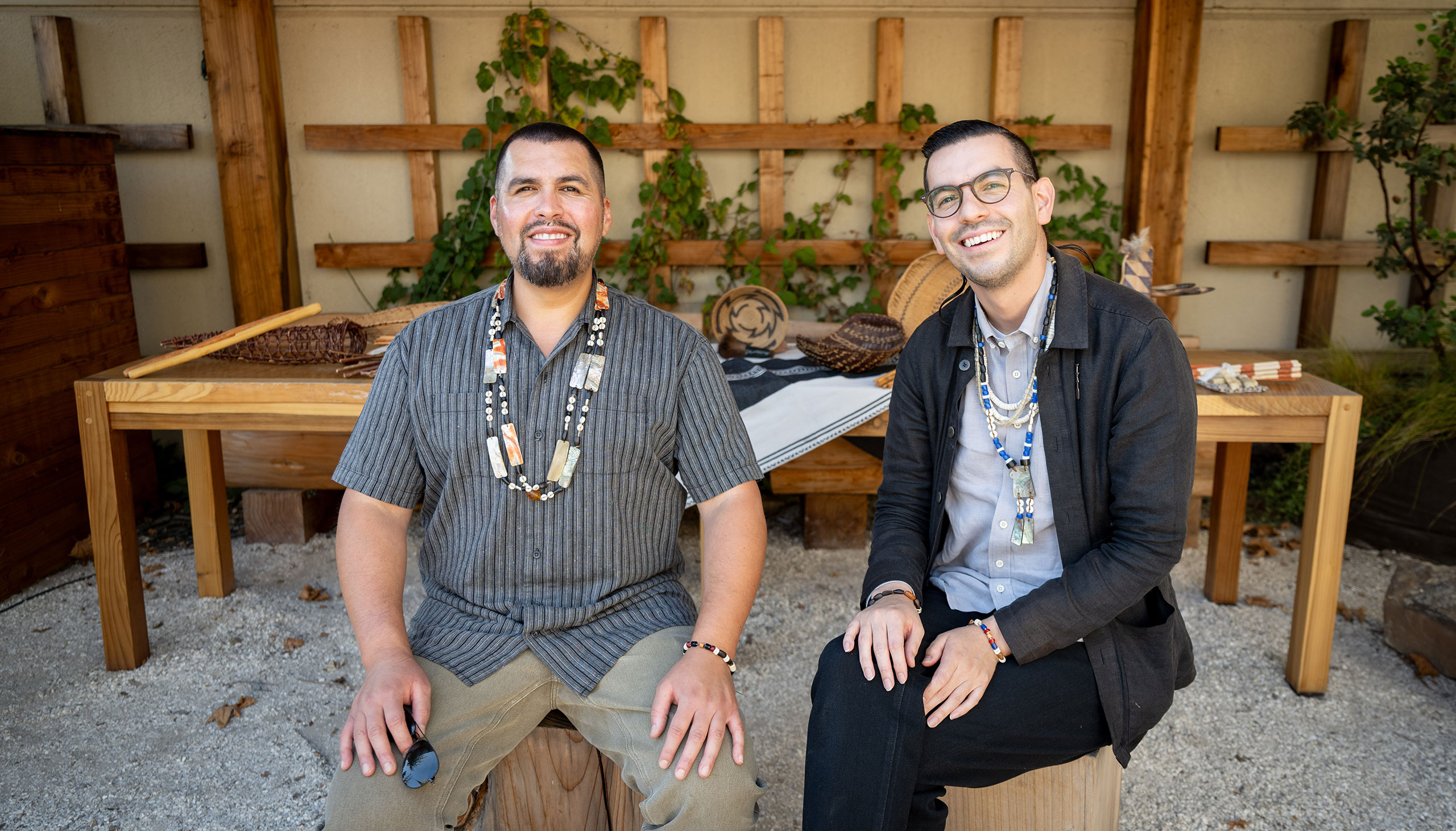 Two people sit on chairs made from tree trunks and smile at the camera. Behind them is a long wooden table that holds basketry and serving utensils.