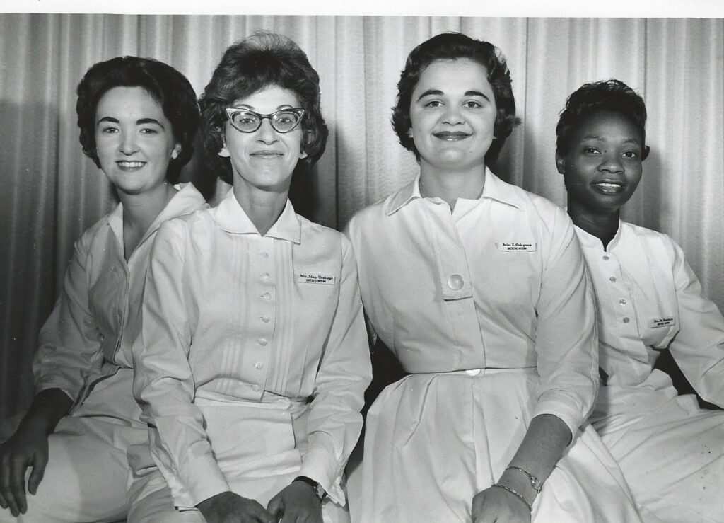 A black and white photo of three white woman and one Black woman, all with 1960s hair and wearing white uniforms.