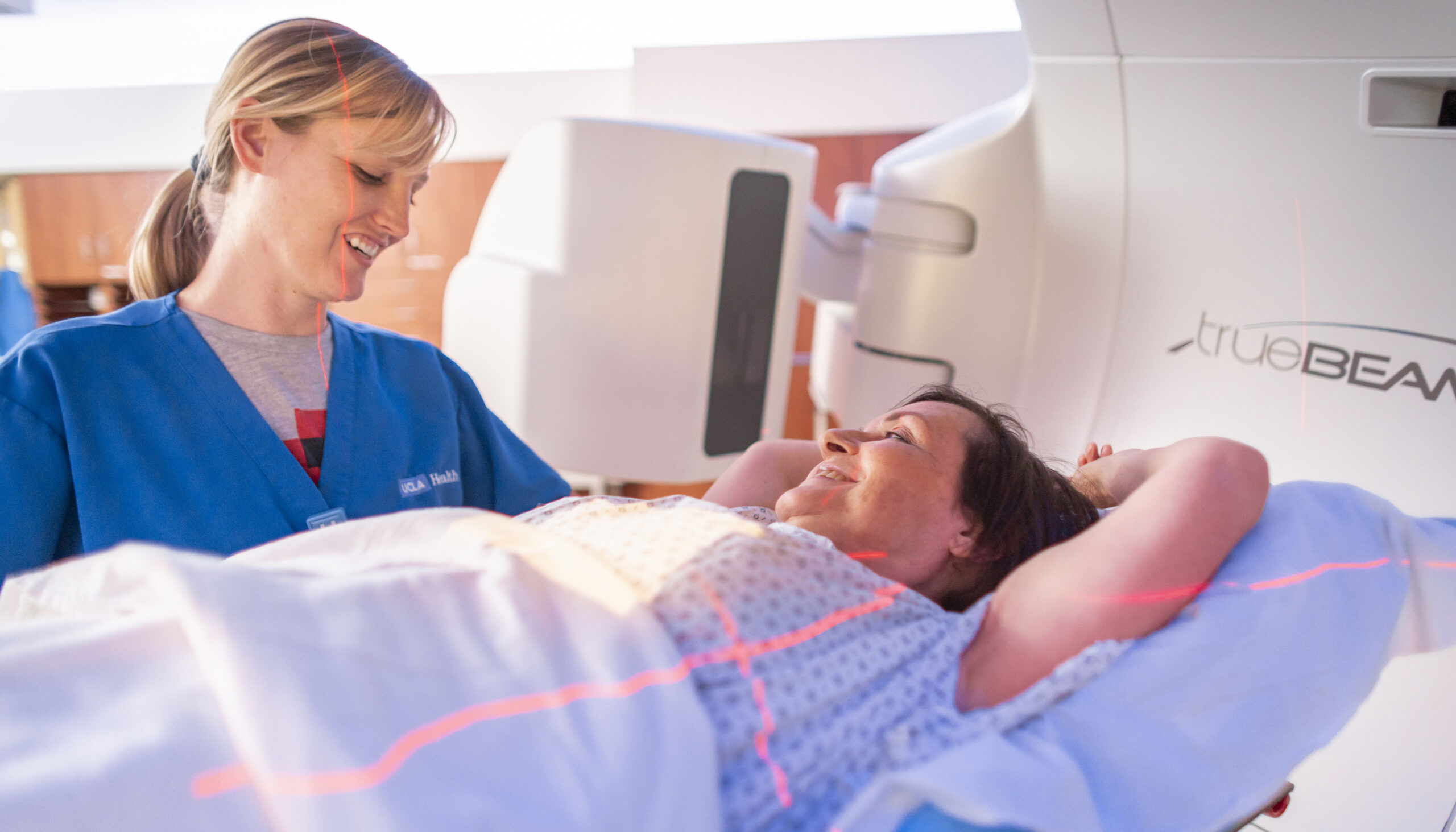 a woman in a blue hospital gown talks to a woman lying on the bed of an imaging scanner