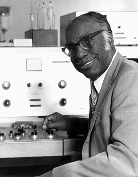 A black-and-white photo of a Black man in glasses and a suitjacket who smiles posing next to lab equipment