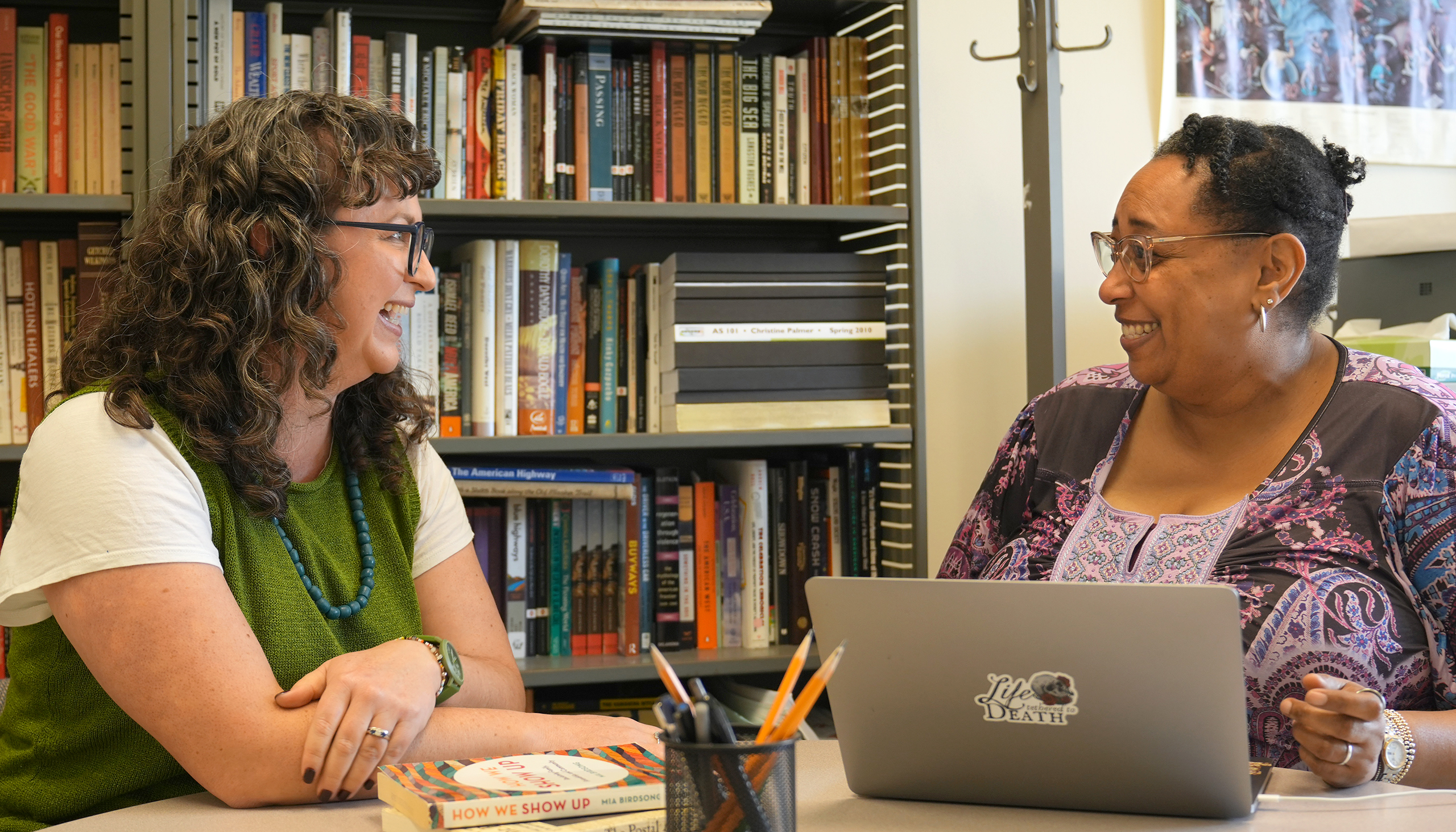 Sarah Gold McBride and Christine Palmer face one another at a desk, smiling at each other, conveying their close friendship