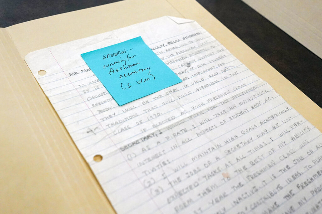 A piece of white lined notebook paper contains a speech that Amy Tan wrote to run for election as freshman class secretary. The letters are in pencil and she attached a blue sticky note to the page later, letting people know that she won.