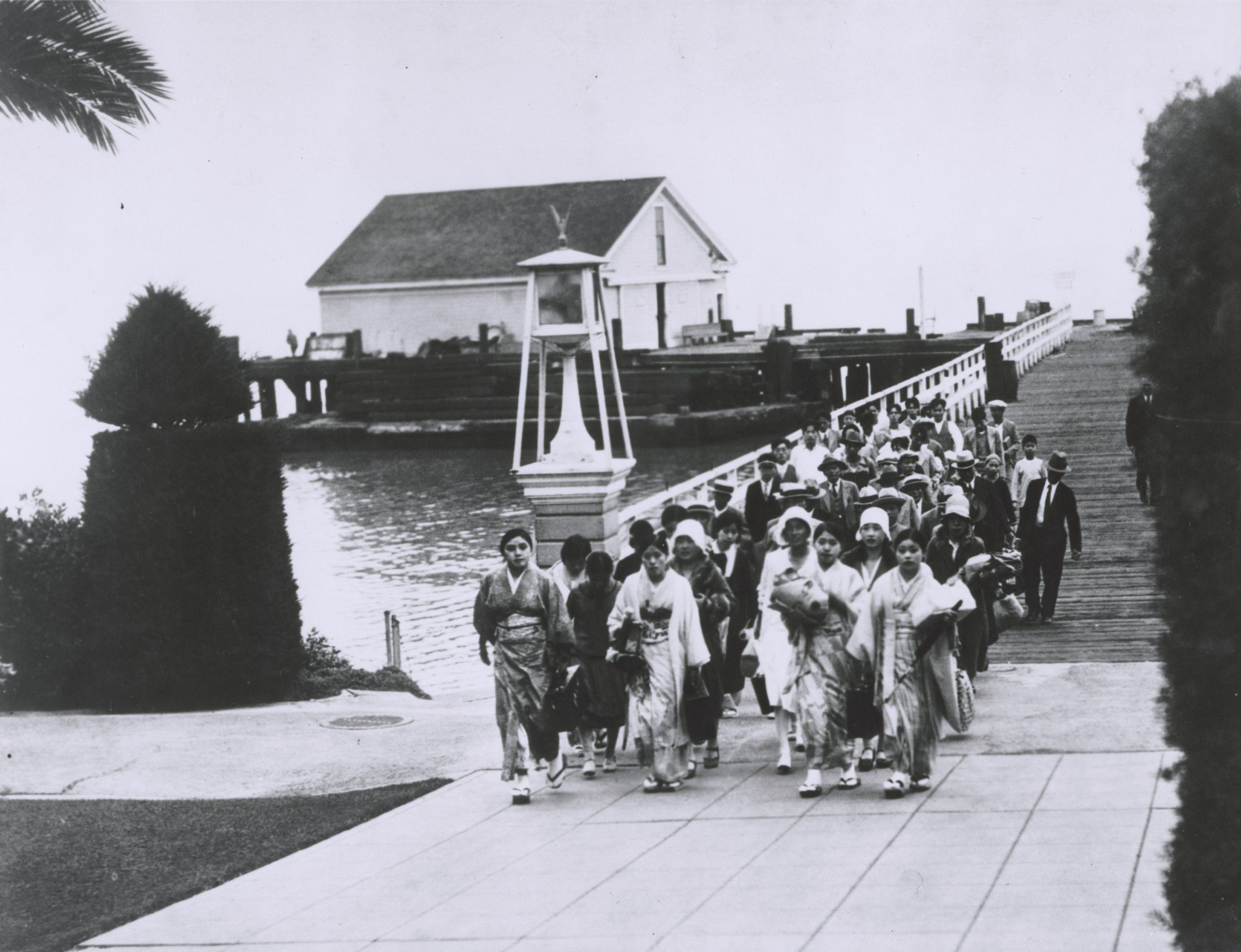 black-and-white photograph of women in traditional dress from East Asia walk across a bridge