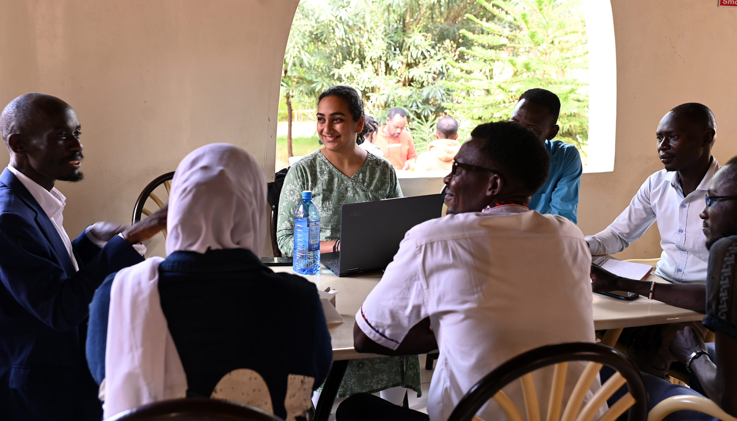 Bhavya Joshi, center, sitting around a table with African colleagues. Behind her, a window opens onto a forested landscape.