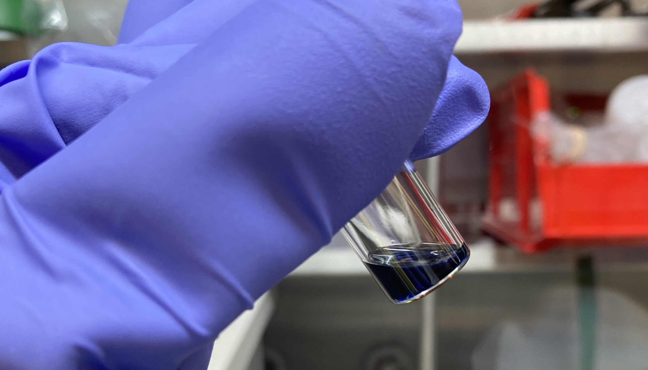 purple-gloved fingers hold a glass vial with black liquid at the bottom