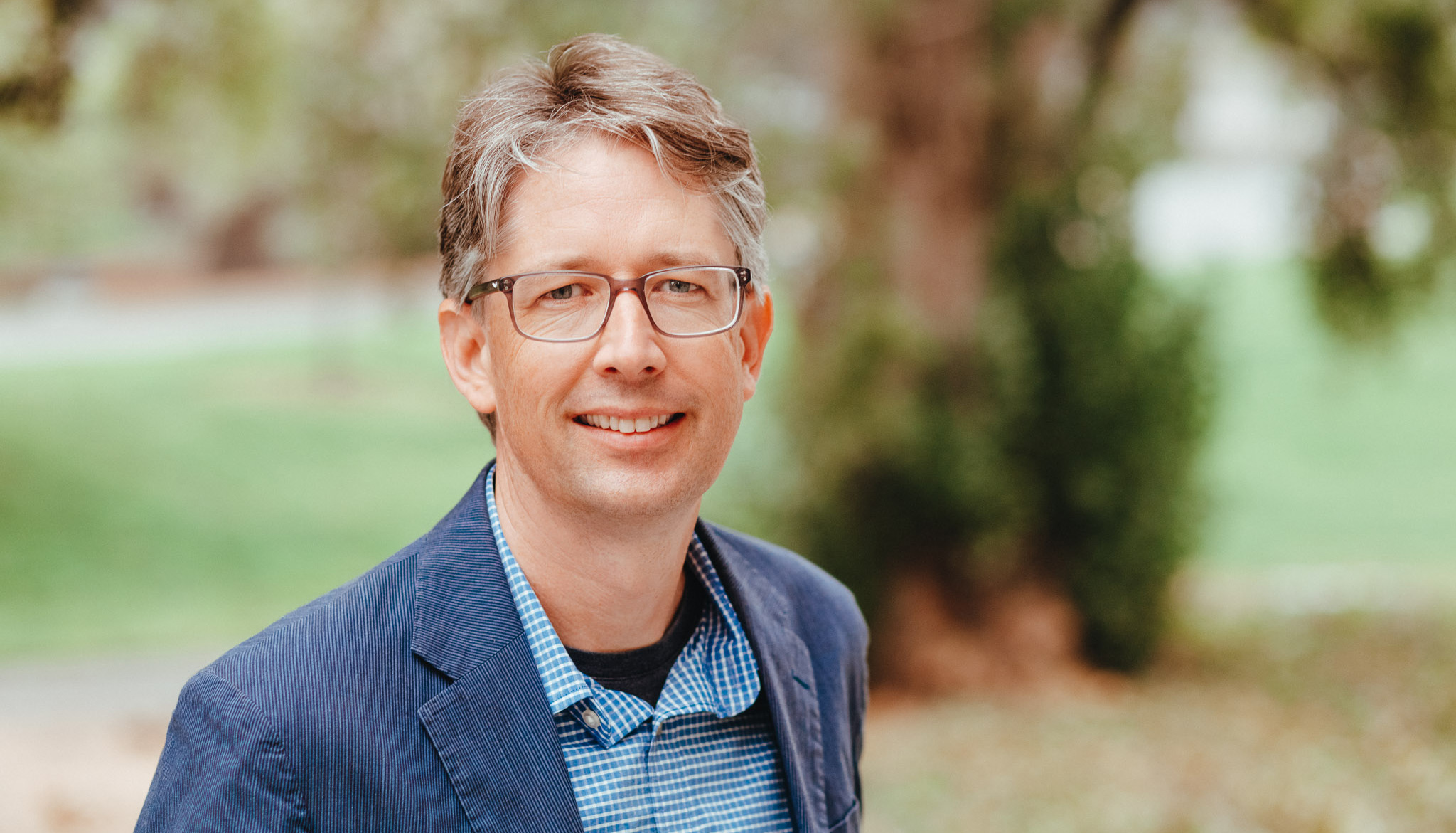 man with blond hair and glasses in a blue shirt and jacket, looking at the camera in an outdoor setting