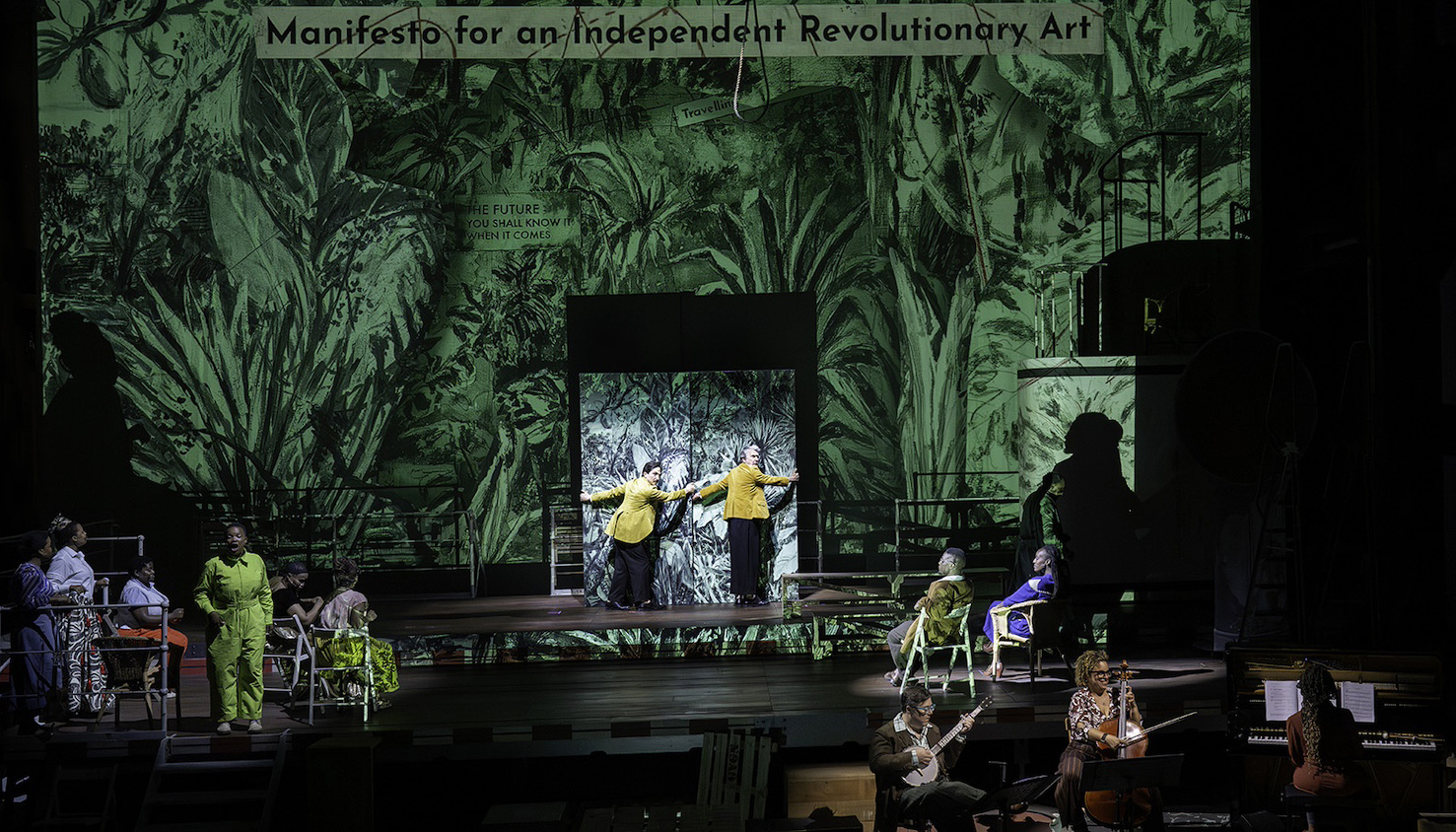 A performance on stage of the chamber opera The Great Yes, The Great No. Two performers attempt to move a panel with an image of green-tinted lush plants on it, which is also projected on a backdrop. Several performers sit in an audience as watch.