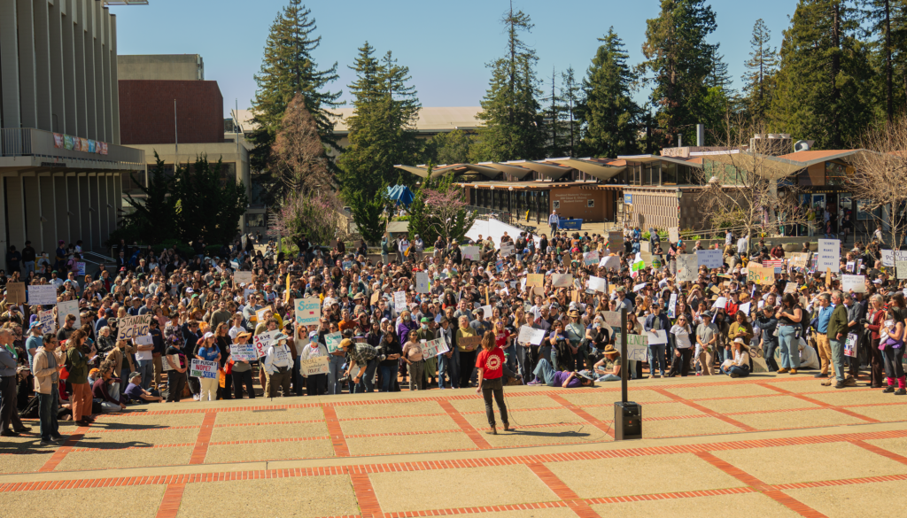 A person in a red shirt addresses a crowd of people holding signs at Sproul Plaza