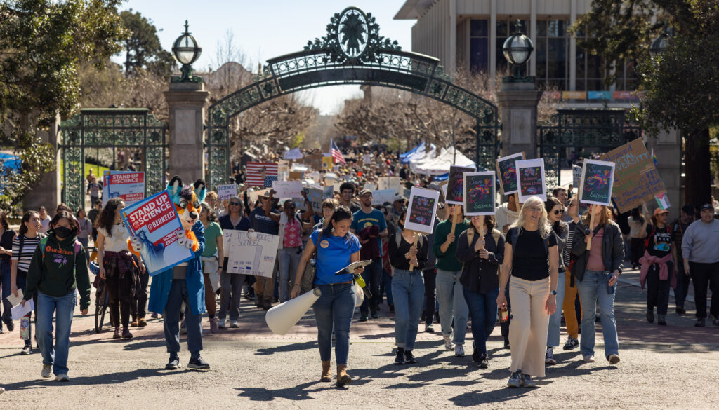 A crowd bearing signs of signs through Sather Gate