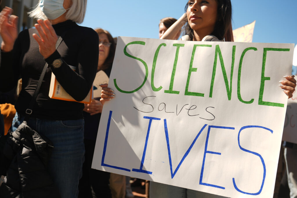 A young woman in a crowd holds a sign reading "Science saves lives"