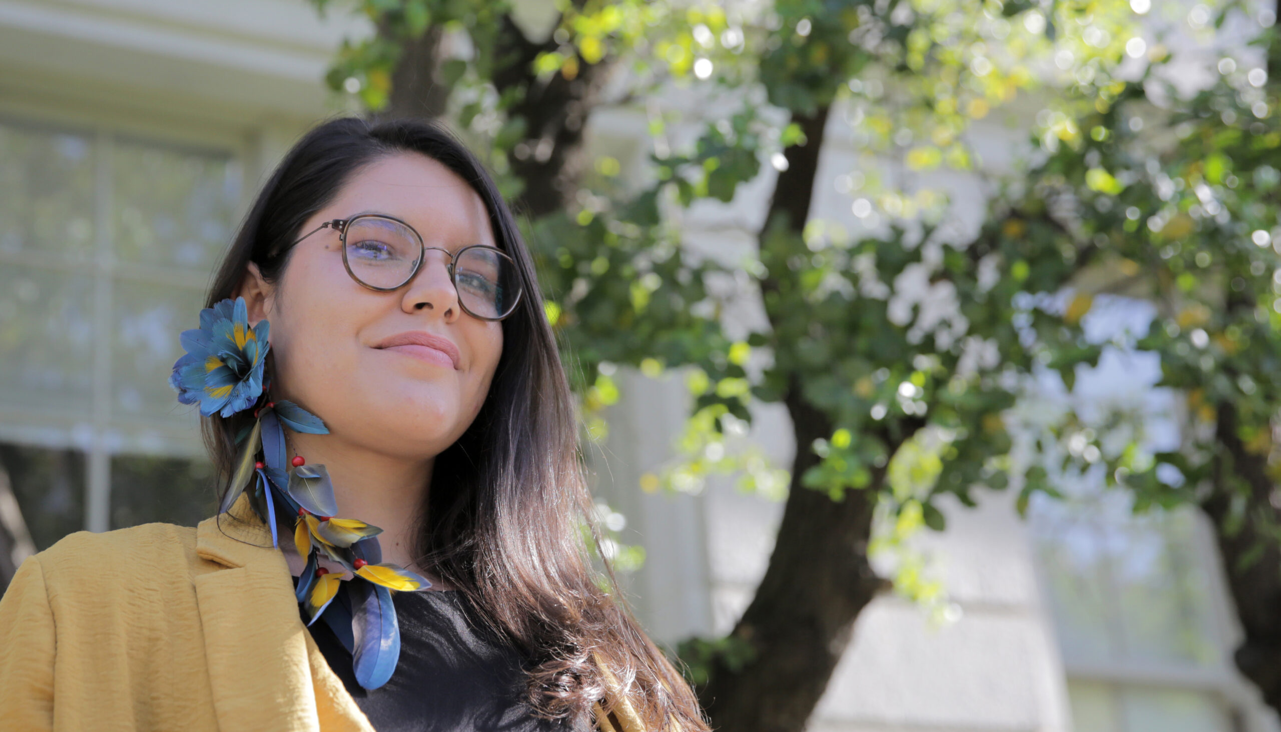 a woman with dark hair and glasses wearing a blue and yellow feathered earring