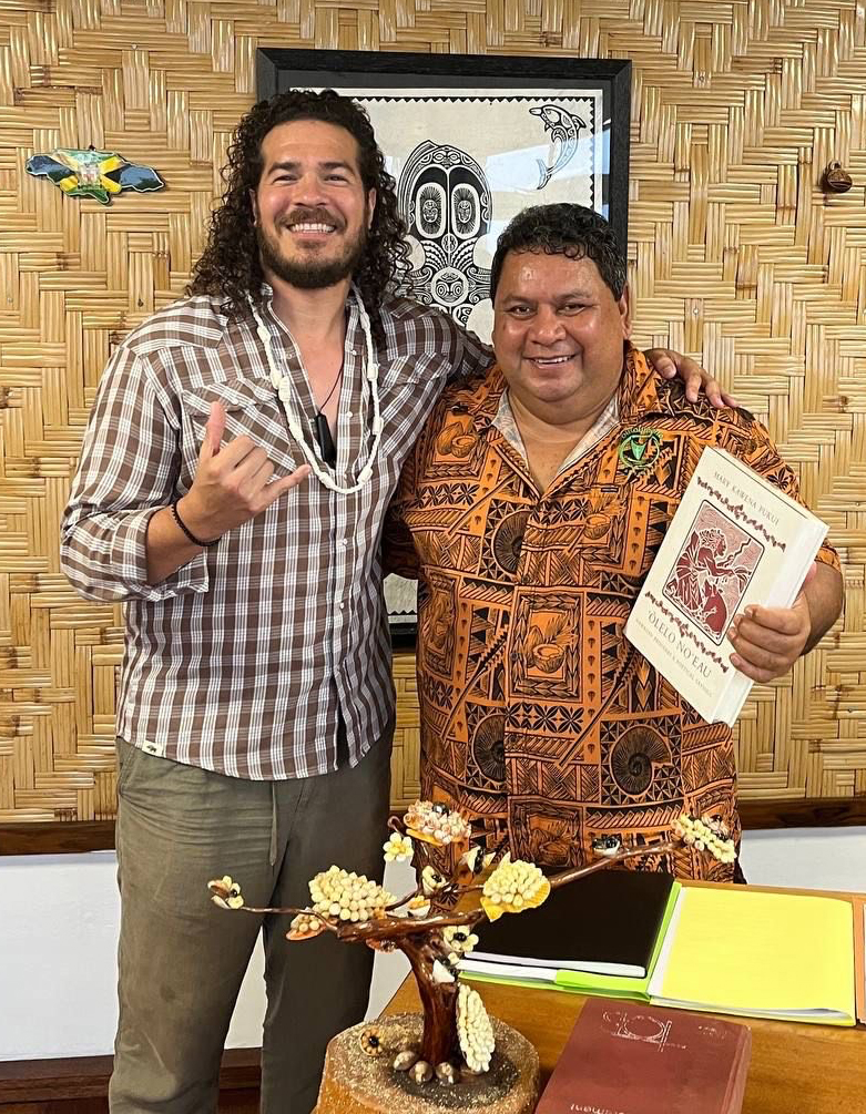 two men in Polynesian shirts hugging, with one making the Love-You hand gesture