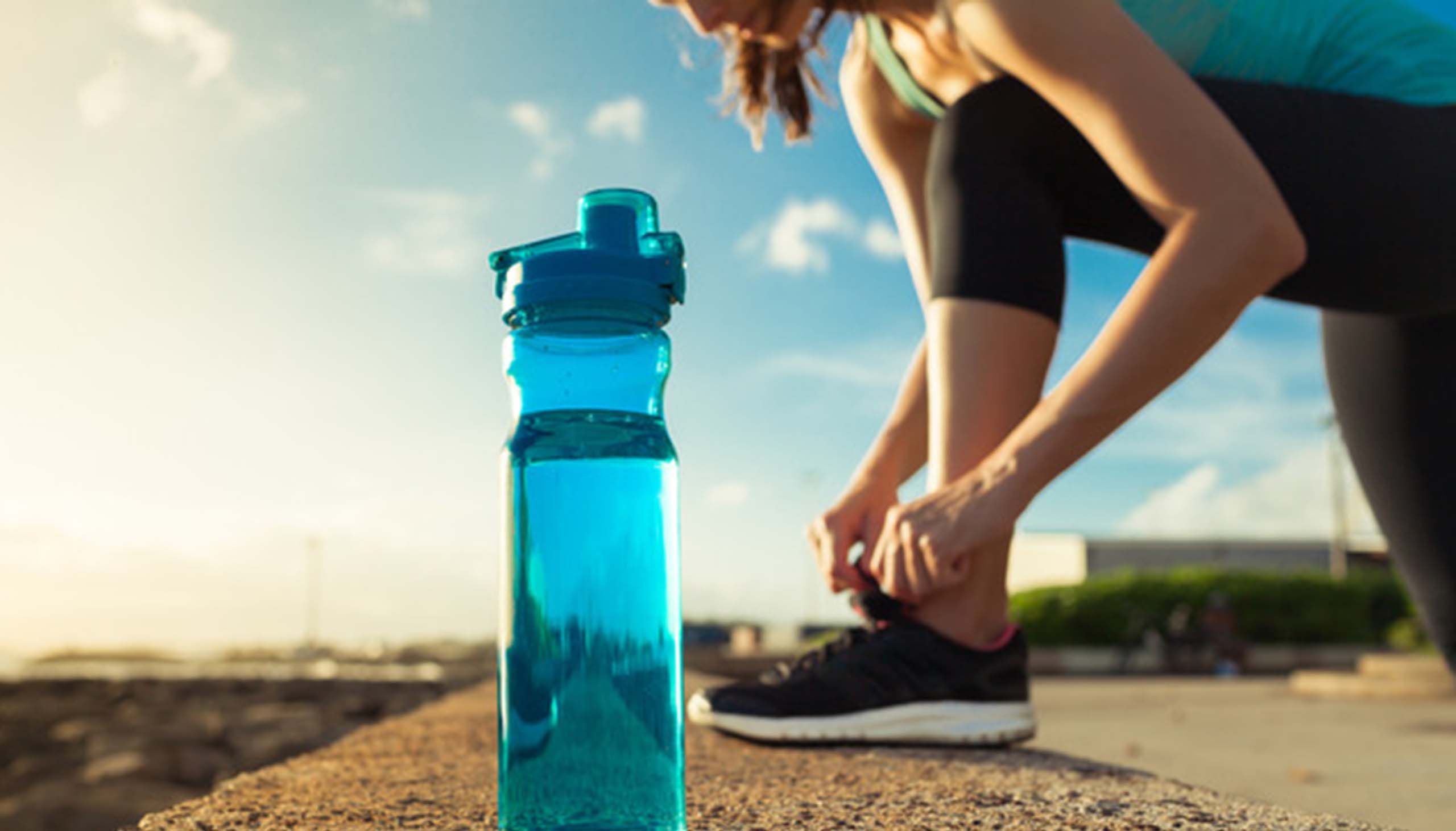 A person bends over to tie their running shoe, with a water bottle in the foreground.