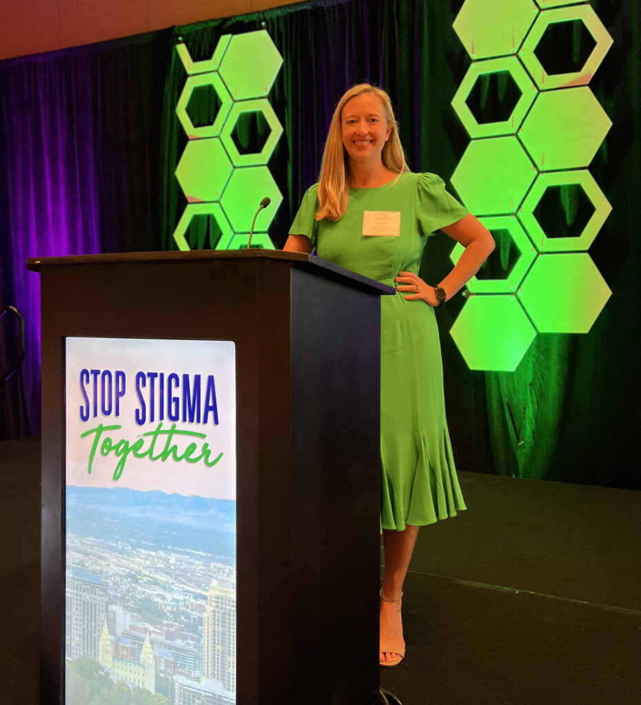 a woman in a green dress standing at a podium holding a sign 'Stop Stigma Together'