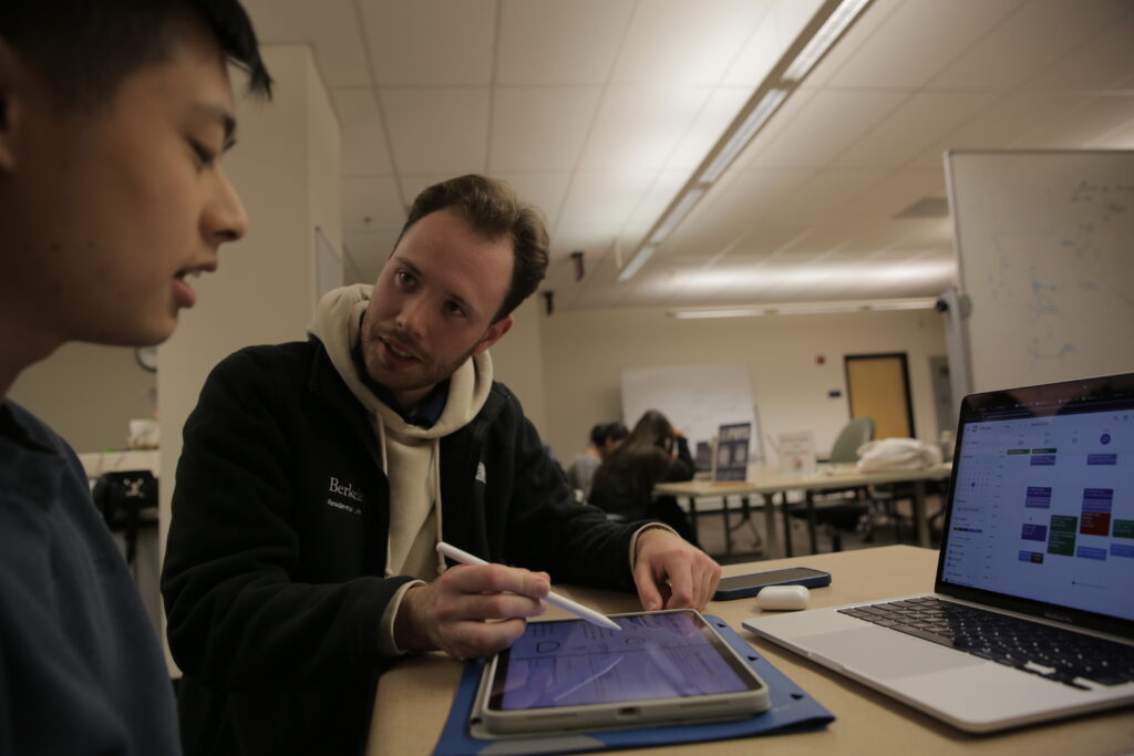 A young man holds stylus and looks at another student as they consider a laptop with a calendar and a tablet that the other student is looking at.