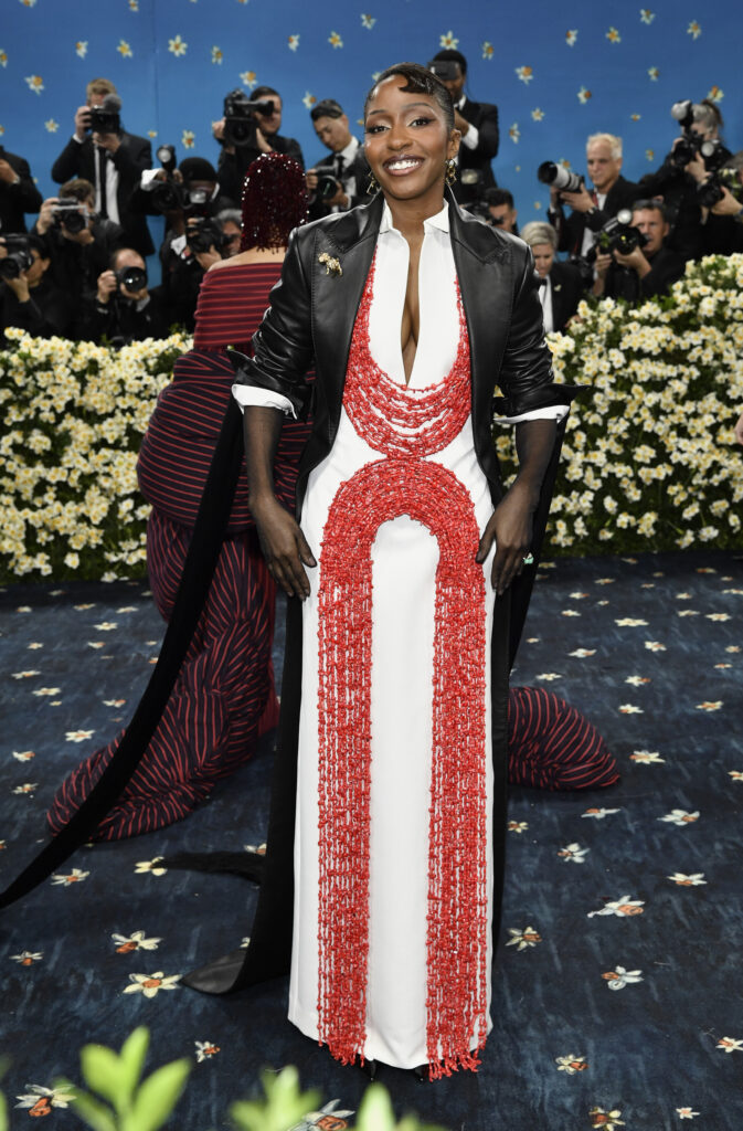 Ayo Edebiri poses wearing a white dress with coral beading in two "U" shapes and a black leather jacket. Her hair is styled with a dollop curl.