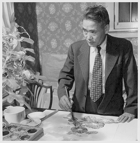 A black and white photo of artist Chiura Obata, a professor at UC Berkeley who was put into a Japanese American internment camp. He is in a suit and tie and painting flowers on a canvas on a table top in Webster Groves, Missouri,. He is looking at a flower arrangement on the table as he paints and holding the brush in his left hand.