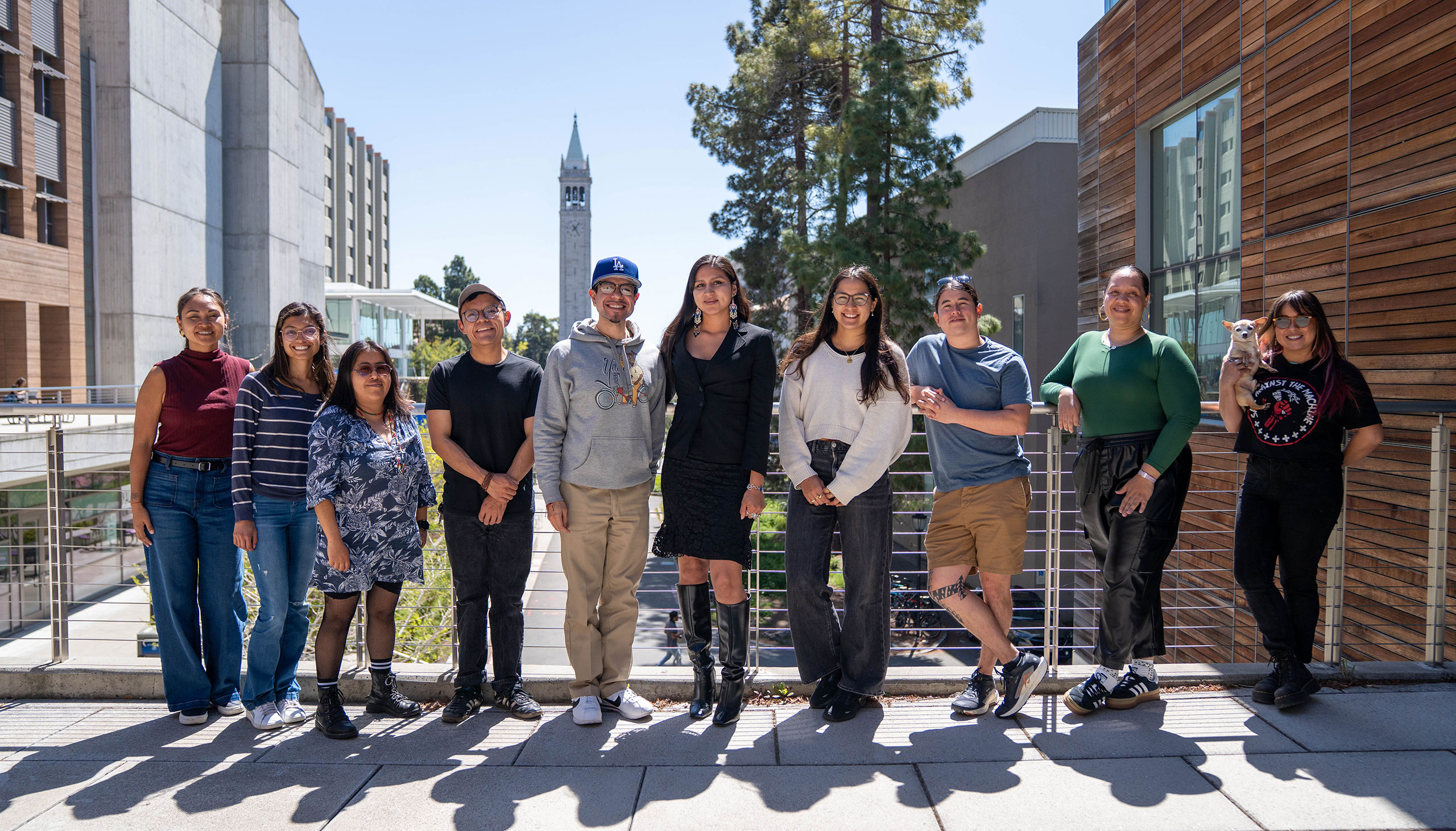 Nine students stand outside on the UC Berkeley campus, with the campanile visible in the background.