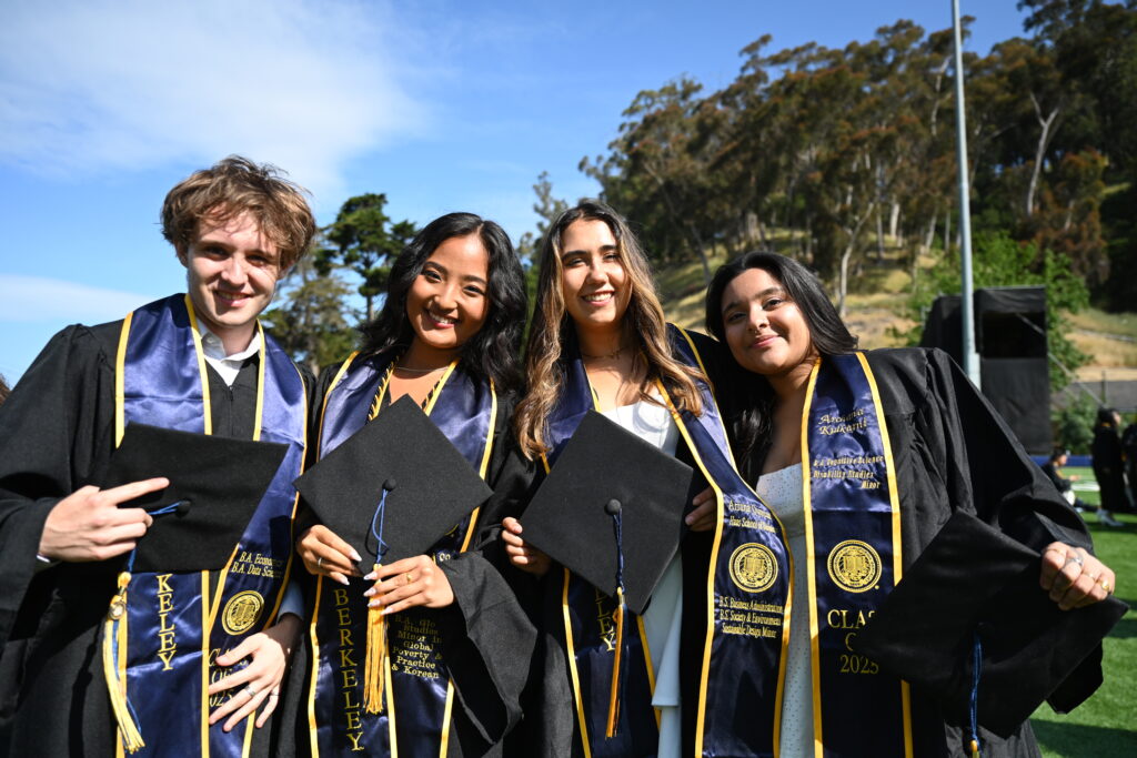 a white man, Korean woman, Latina woman, and Indian woman smile at the camera and hold their graduation caps, wearing embroidered blue Berkeley stoles
