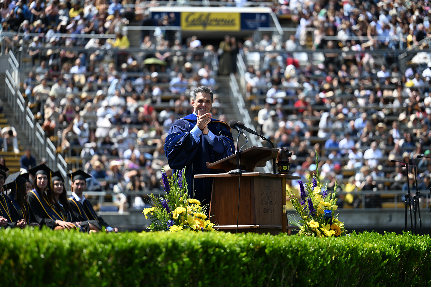 At the 2025 campuswide commencement, Daniel Lubetsky, the founder of KIND bars, applauds grads from the podium. He is wearing regalia.