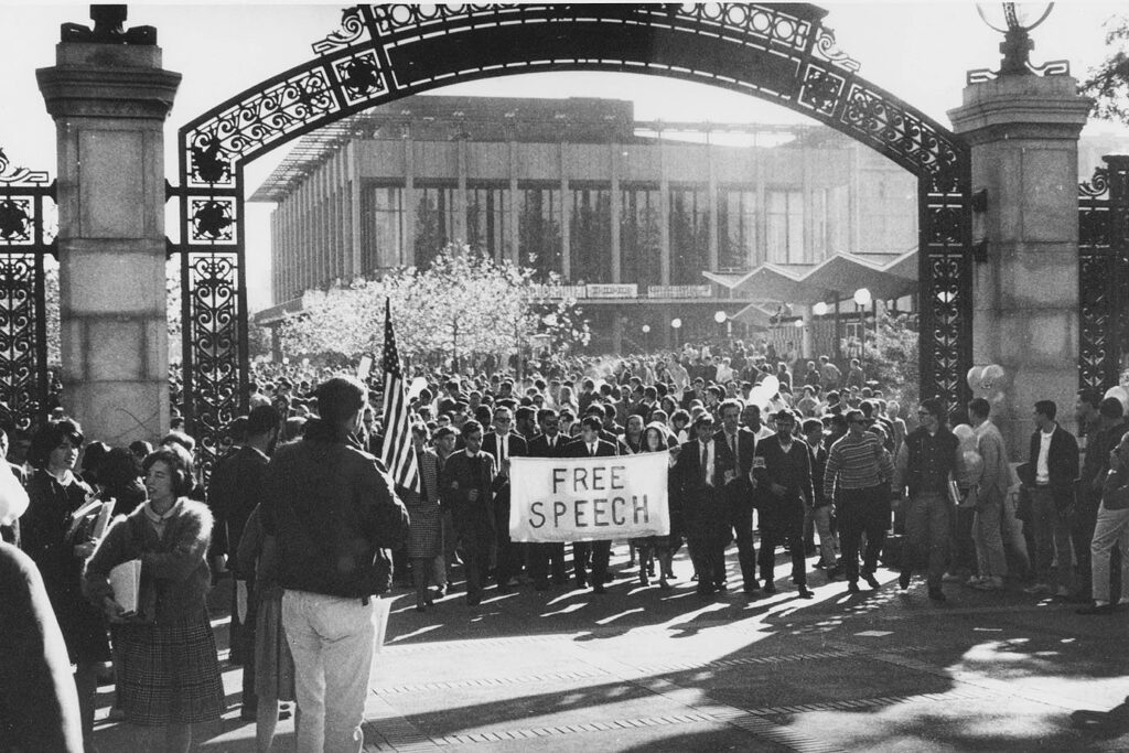 In a black & white photo, protesters in 1964 march through UC Berkeley's landmark Sather Gate carrying a banner that reads "Free Speech."