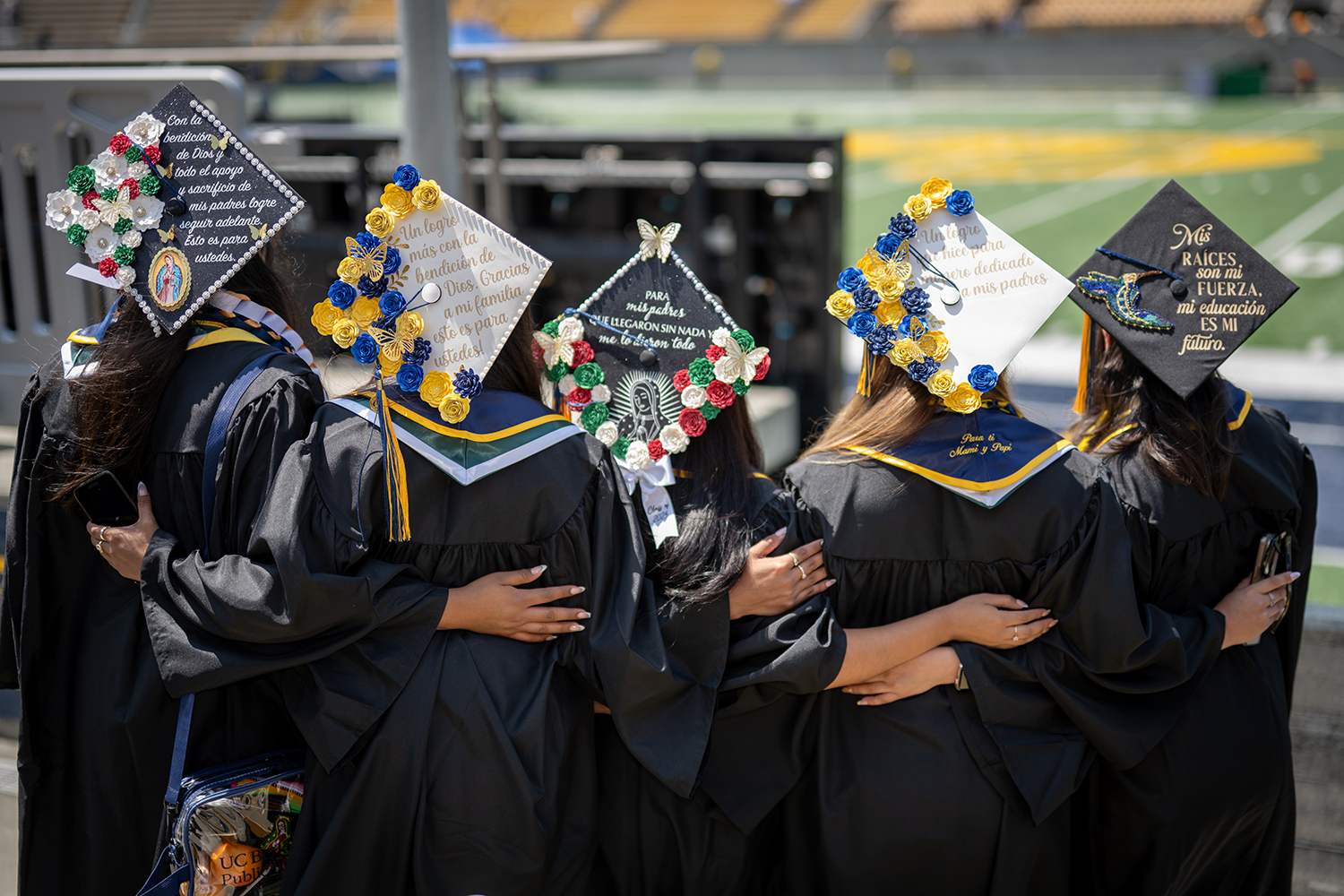 Five friends with individually decorated mortarboards stand with their backs to the camera with their arms wrapped around each other.