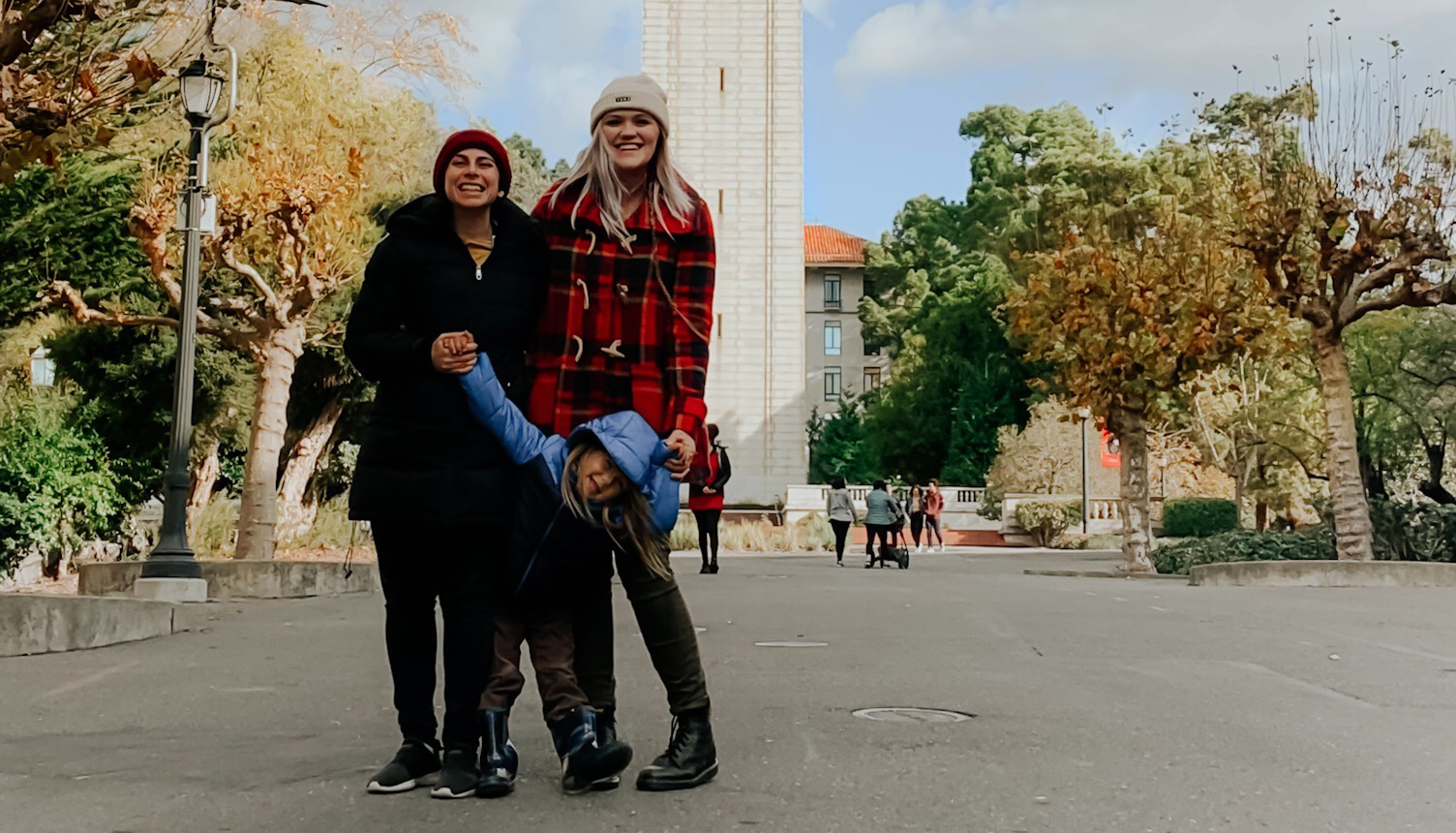 Emily and Yasmin stands smiling with their 4-year-old kid, River, in front of the Campanile in 2019.