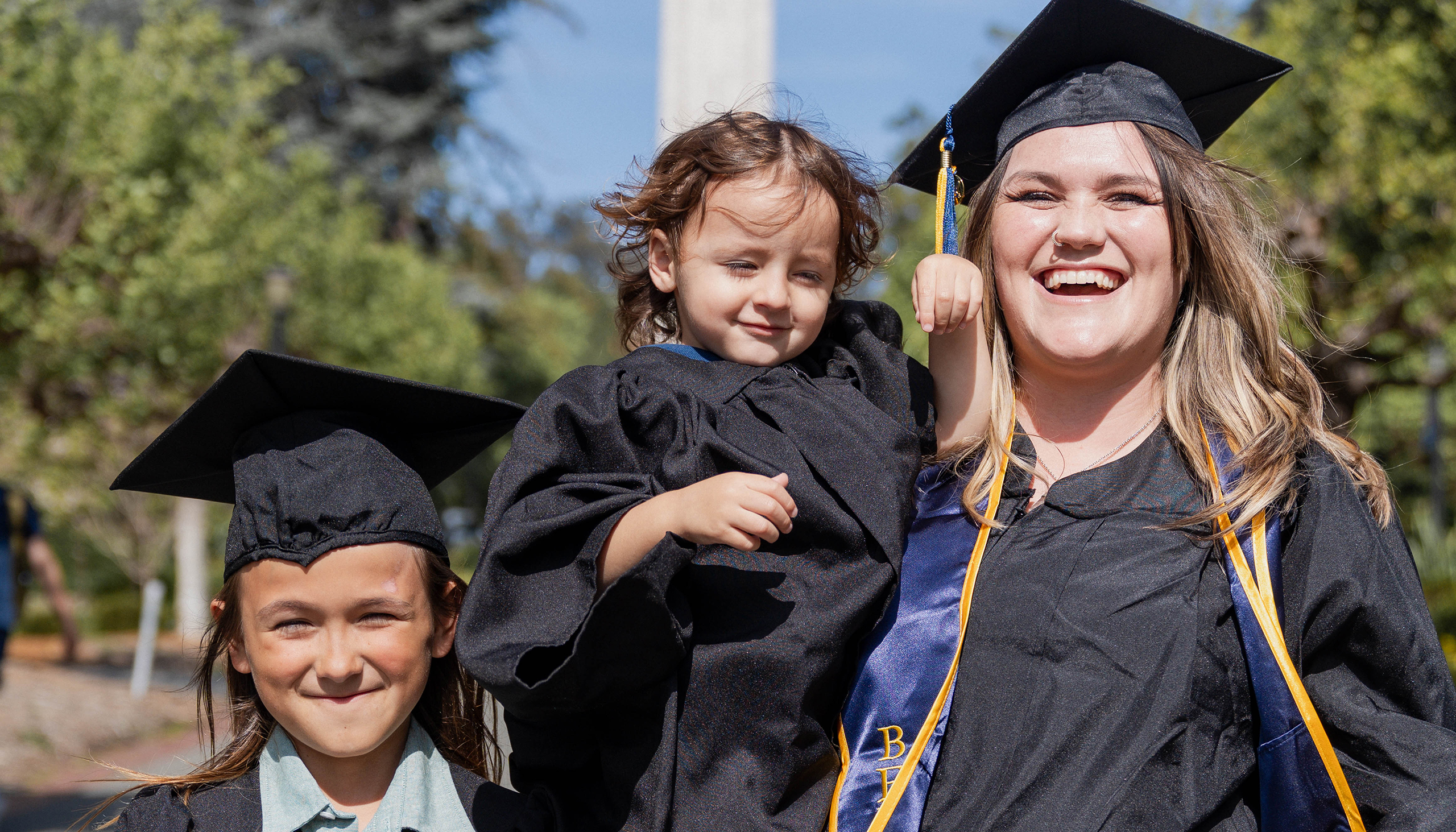 Emily Thompson, clad in a cap and gown, holds her 2-year-old, Caspian, also wearing a small gown, while her other kid, River, stands next to her. She's smiling widely.