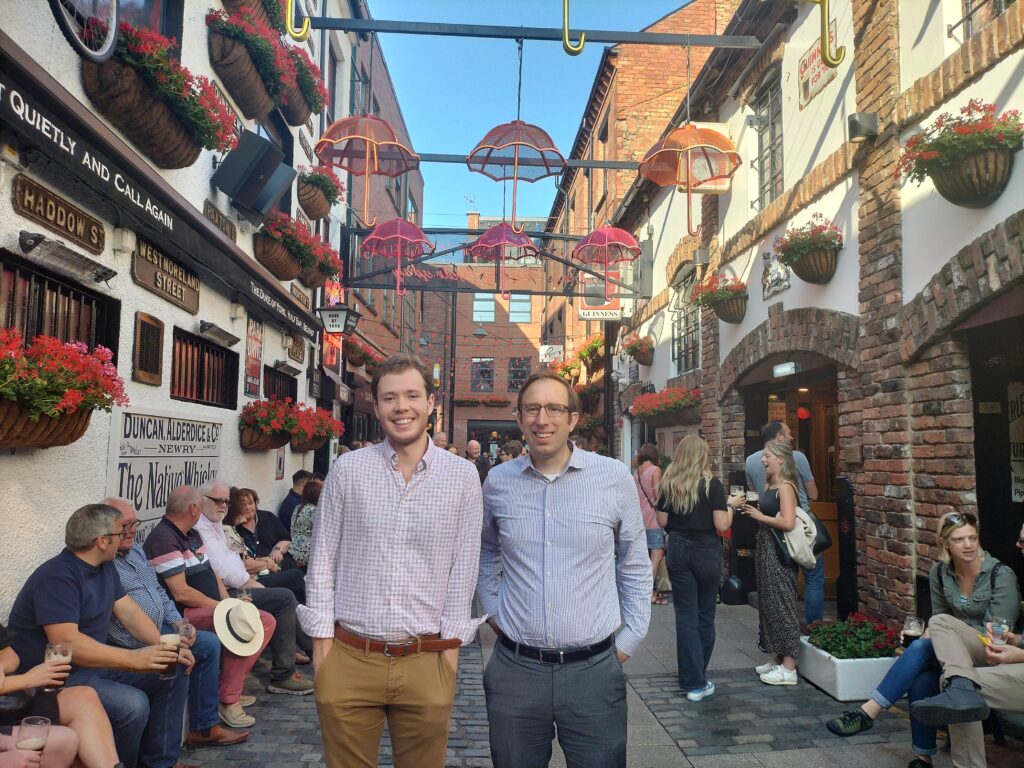 two men in collared shirts stand in a street with whimsical umbrella decorations overhead.