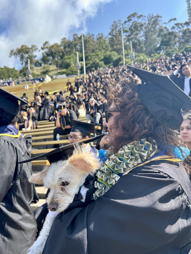 The profile of a woman in graduation regalia, wearing a lei with dollar bills and holding a cream dog who is also wearing a graduation cap. Behind her, students sit in the stands.