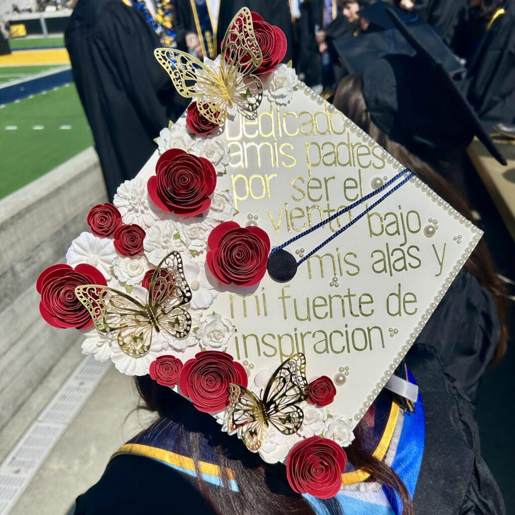 a mortarboard with a white background. On the left side, there are red and white paper flowers as well as golden butterfly cutouts. The right edge is lined by small pearls. In gold lettering, it reads, Dedicado a mis padres por ser el viento bajo mis alas y mi fuente de inspiracion.