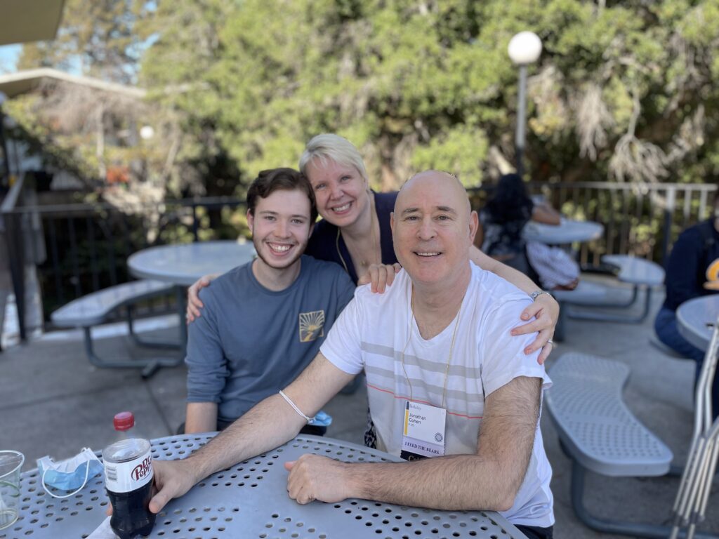 A young man smiles at a plastic table with his parents. The mother has her arms around the father and son, who are seated. There is a blue mask on the table and on the father's arm, and the dad wears a name tag.