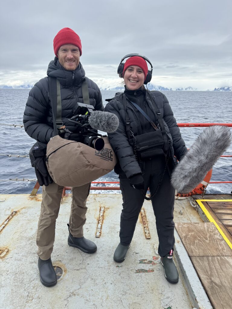 two people wearing rainboots, red hats, and black jackets on board a boat with a gray sea in the background. The man is holding a camera and the woman has a giant fuzzy microphone and headphones on.