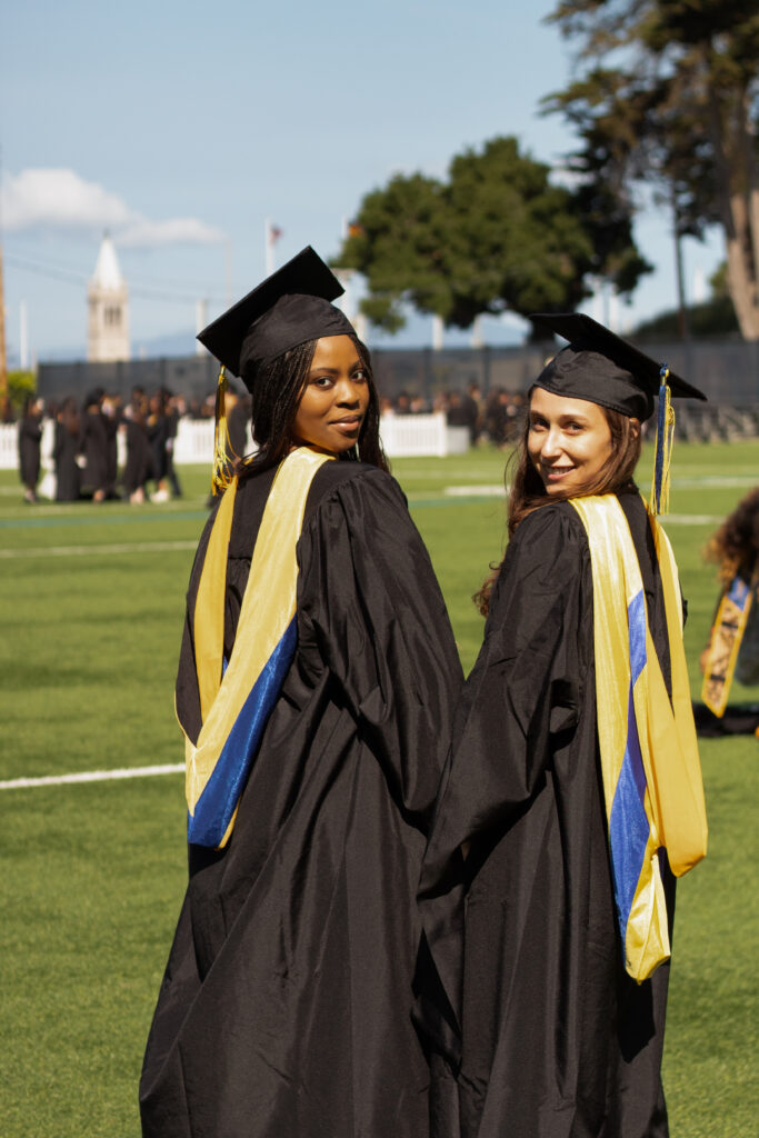 A Black woman and white woman look over their shoulders and smile. They are dressed in graduation regalia and wearing blue and gold hoods that show they are receiving master's degrees.