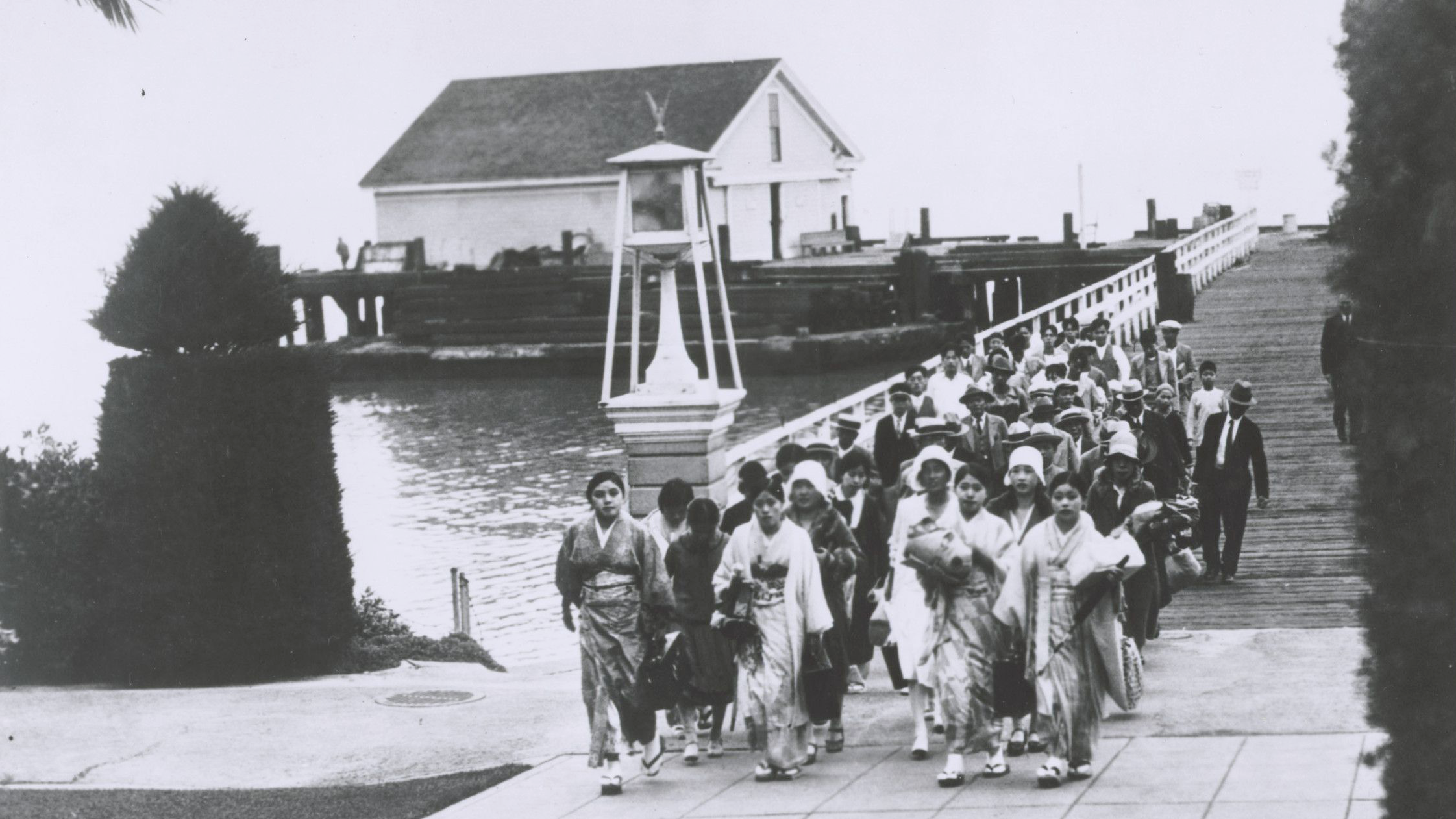 black-and-white photograph of women in traditional dress from East Asia walk across a bridge toward Angel Island in the early 1900s