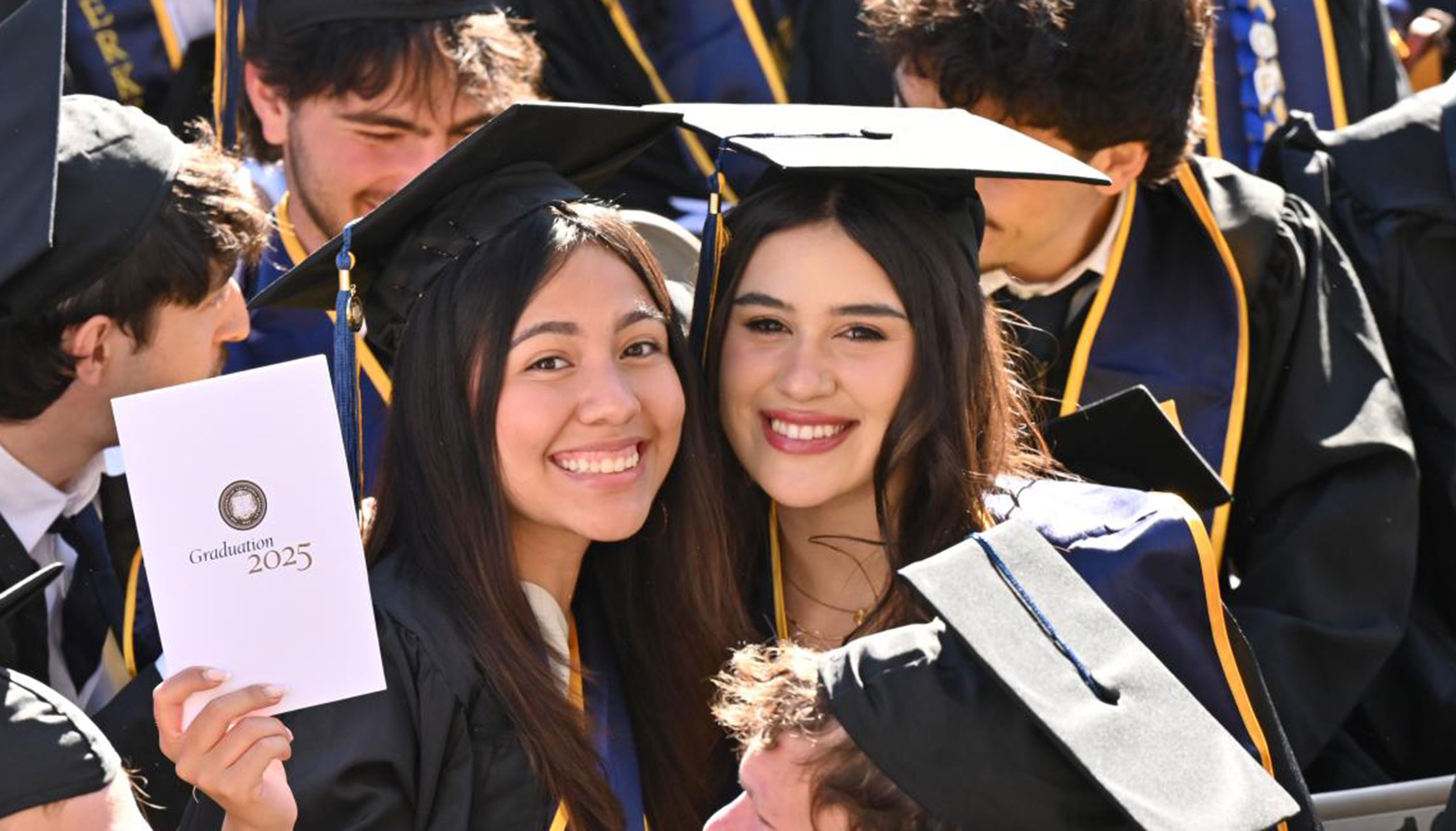 Two people wearing caps and gowns stand together and smile amidst a crowd of other graduates.
