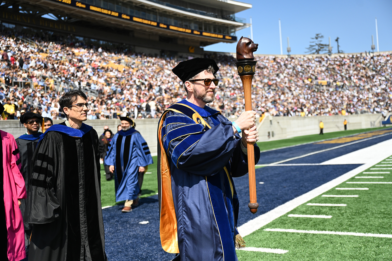 Wearing regalia, Ben Hermalin, the campus's executive vice chancellor and provost, carries the campus's mace into the football stadium in the 2025 campuswide commencement procession. The mace is a highly ornamental stick carved of wood with a bear's head at the top.