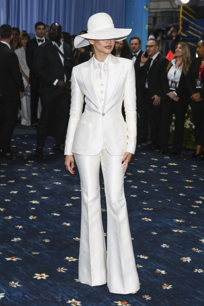 Zendaya on the blue carpet, wearing a white suit with flared legs and a wide-brimmed hat that covers her eyes
