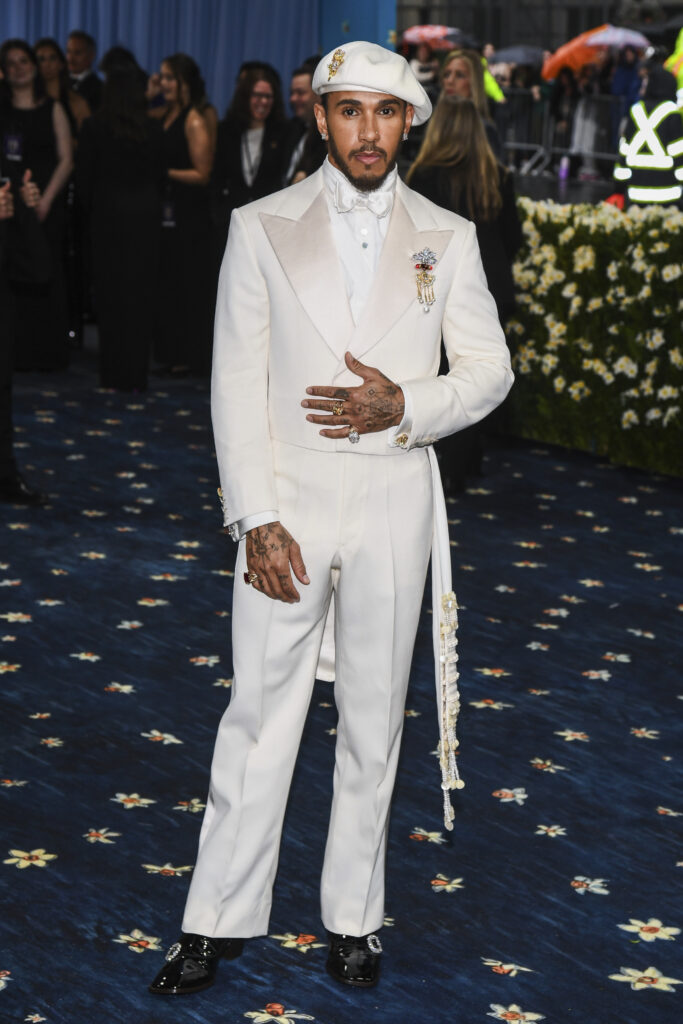 Lewis Hamilton, wearing a white newsboy cap, cream suit, jewelry, and a cowrie shell accessory piece