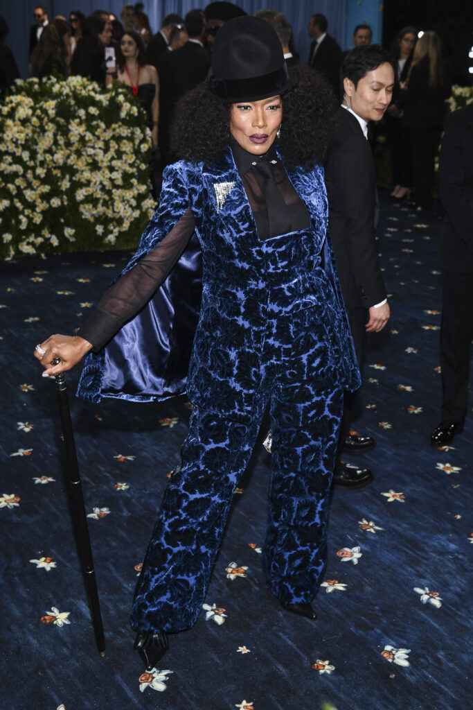 Angela Bassett on a blue carpet. She holds a cane in one hand and is posing in a blue velvet suit and wearing a tall black velvet hat