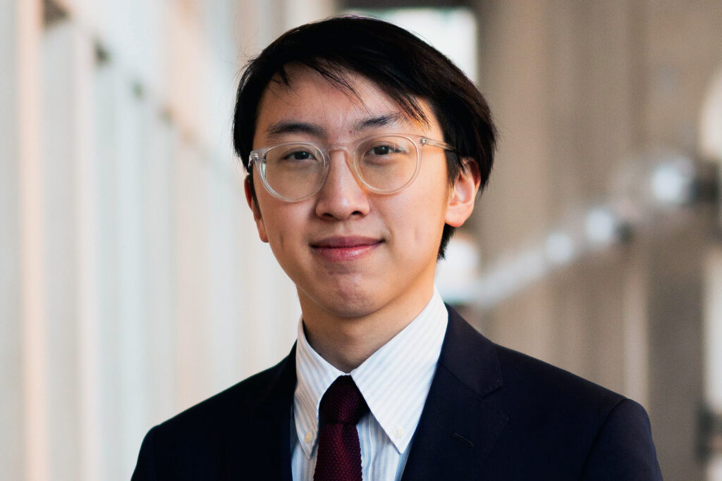 informal headshot of Nathan Tang, wearing a dark suit and a dark tie, against a geometric architectural background