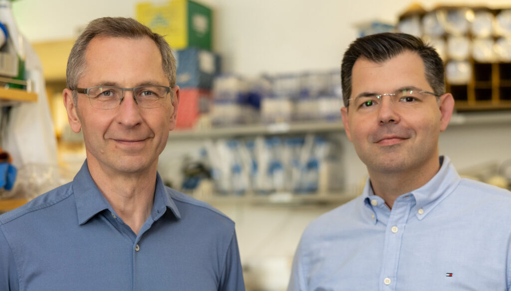 two men in blue shirts smiling into the camera, with lab equipment in the background