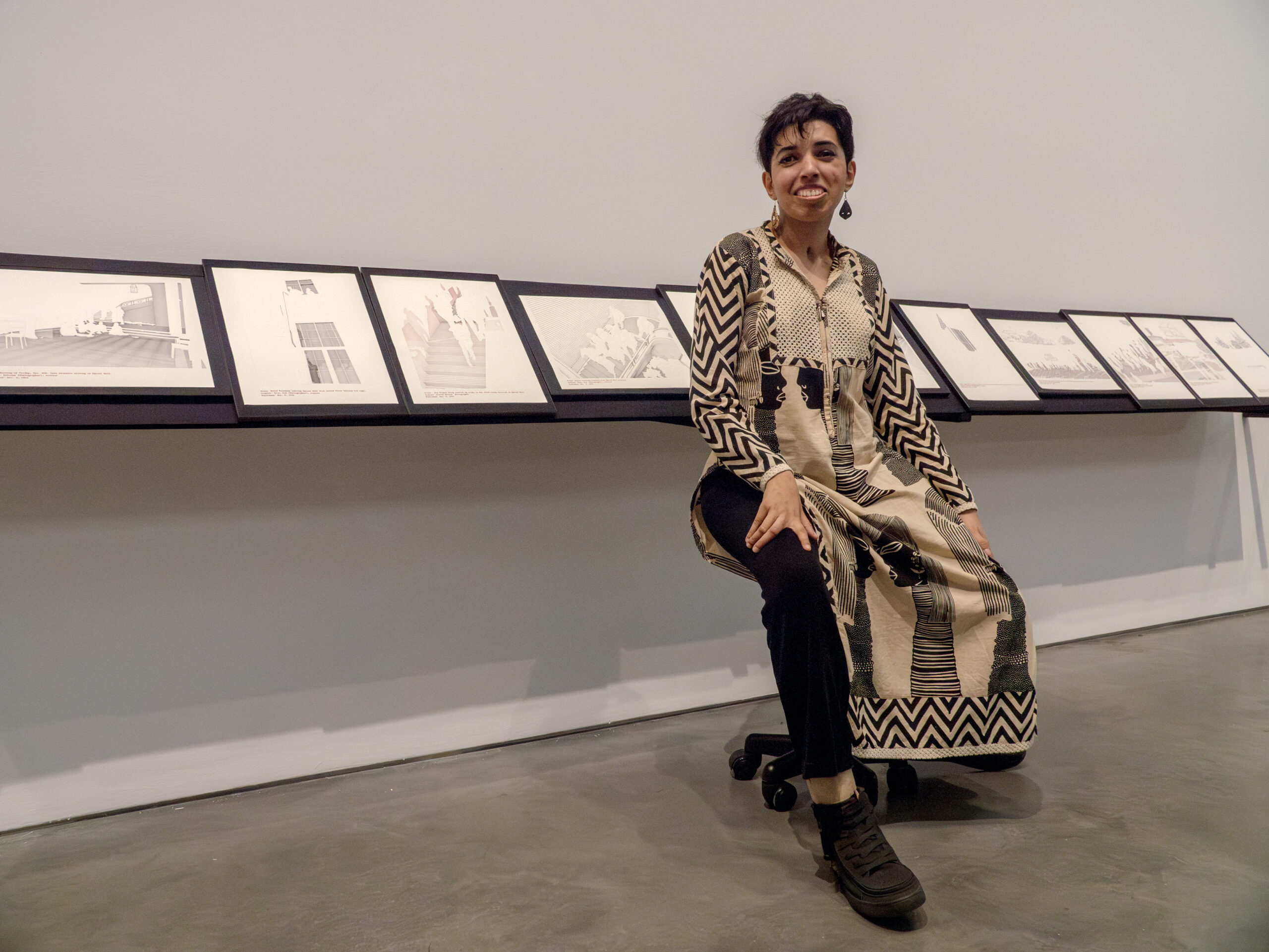 Priyanka D’Souza, pictured sitting on one of the wheeled stools installed as part of her MFA exhibition, before various works created for the exhibition mounted on a shelf viewable at seated height.